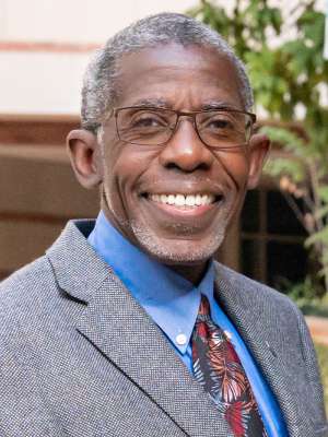 Headshot of Doctor Dale Abel in a grey suit, wearing glasses and standing in front of a brick building with trees to the right.