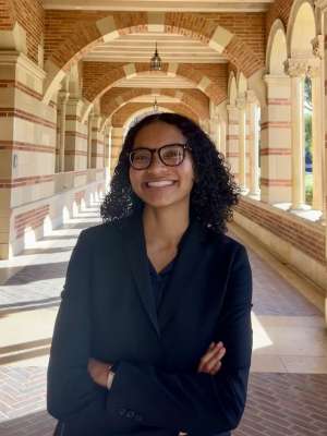 A woman with dark brown curly hair and glasses smiling, standing underneath the sunlit arches