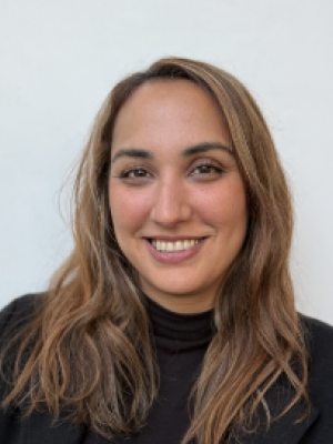 Shandiz Kamrava with light brown hair wearing a black top and jacket, smiling in front of a plain background.