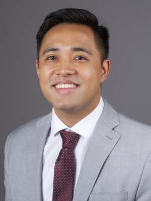 Smiling man wearing gray suit and maroon tie against a neutral gray background.