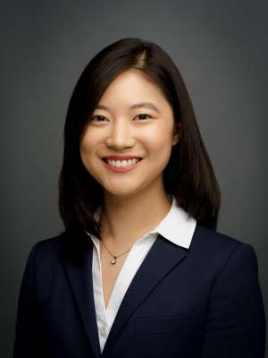 A woman with black hair, wearing a navy suit jacket with a white shirt and necklace