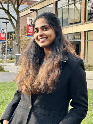 A person with long hair wearing a black coat stands outdoors near a building.