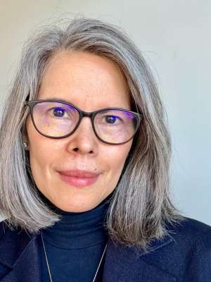 Adult woman with gray hair, glasses, navy blazer and black turtleneck against light background.