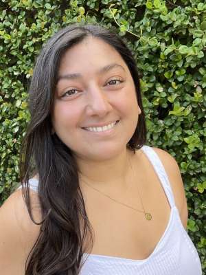 A woman with black hair, wearing a white top and necklace, smiling in front of a leafy background