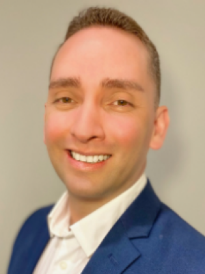John Avila in a blue suit jacket and white shirt smiling in front of a plain background.
