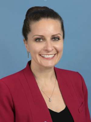 Professional headshot of a woman in a red blazer against a blue background.