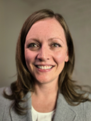 Danielle Spitzmiller with shoulder-length brown hair wearing a light gray blazer and white top, smiling against a plain background.