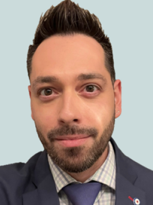 Robert with short dark hair and beard wearing a suit, checkered shirt, and tie, smiling in front of a plain background.
