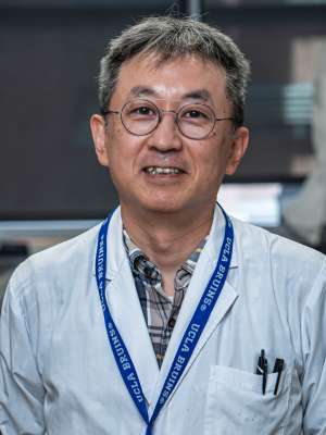 Kenuske Tsushima wearing glasses, flannel shirt, white lab coat, and a lanyard saying UCLA BRUINS. background is blurry.