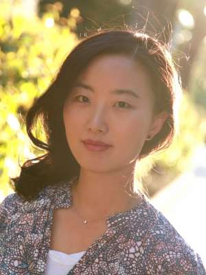 A woman with long black hair, wearing a floral shirt and necklace, posing in front of a sunny forested background