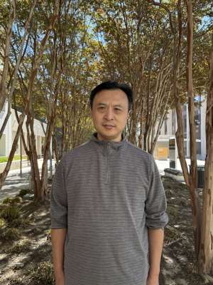 Ke Wang wearing a grey long sleeve shirt with the sleeves rolled up standing in front of some trees