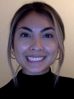 A woman with blonde hair, wearing her hair up with a black sweater and earrings, smiling in front of a blank background