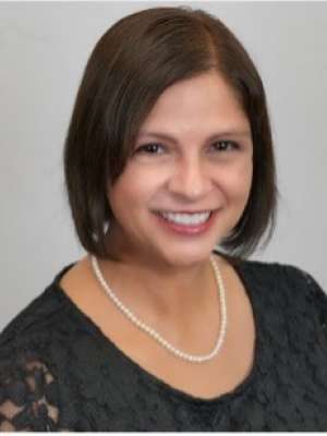 A woman with straight brown hair, wearing a black dress with a necklace, smiling in front of a blank background