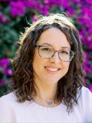 A woman with curly hair wearing a light shirt, surrounded by purple flowers.