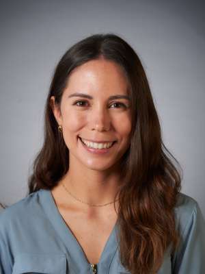 A woman with brown wavy hair, wearing a muted blue shirt, smiling in front of a blank grey background