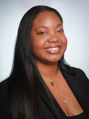 A woman with straightened black hair, wearing a black suit coat and necklace, smiling in front of a grey background