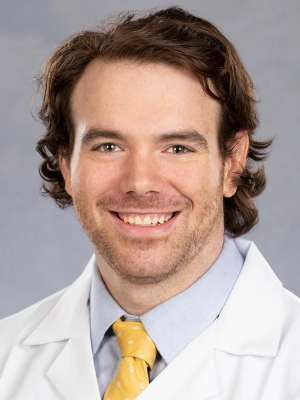 Smiling man in white lab coat, blue shirt and yellow tie.