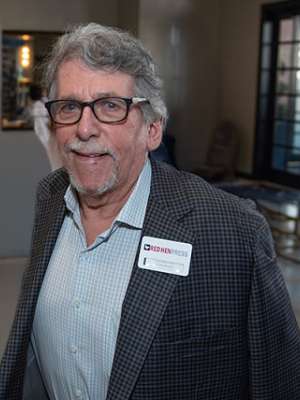 Gary Edelstone in a checkered blazer wearing a name badge, walking in a well-lit indoor space.