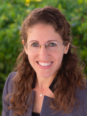 A woman with curly hair wearing a blazer, standing outdoors with greenery in the background.