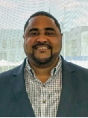 A man with short black hair and a goatee, wearing a blazer and collared shirt, smiling in front of a building window