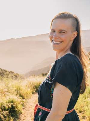 A woman stands on a trail with mountains and sunlight in the background.