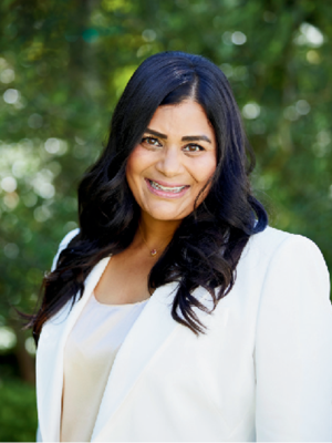 Amita Ramesh with long, wavy hair wears a white blazer against a green background.