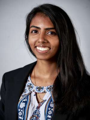 A woman with black hair, wearing a suit coat with a floral shirt, smiling in front of a blank grey background