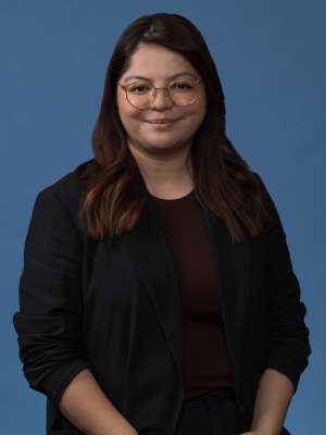 Smiling person with glasses wearing a black blazer against a plain blue backdrop