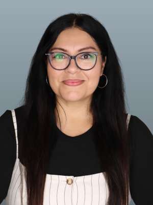 Woman with long dark hair and glasses smiling in front of gray background.