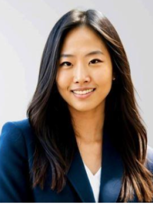 A woman in a dark blazer with long hair, standing against a light background.