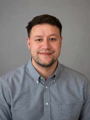 Daniel Cortez - Smiling man in gray button-down shirt against plain gray background.