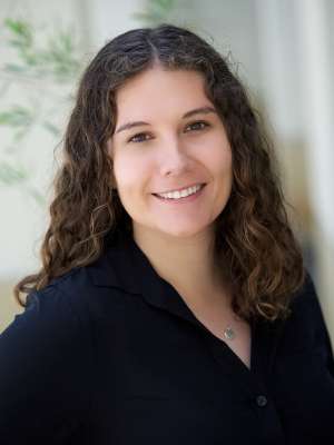 A woman with long brown curly hair, wearing a black shirt, smiling in front of a curtained background