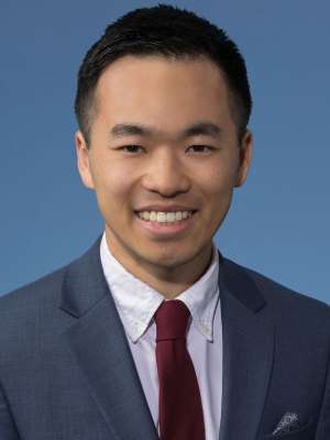 Professional headshot of a smiling person in suit and red tie on blue background.