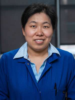 Yuan Zhang wearing a blue buttoned shirt with a blue lab coat. Background is blurry.