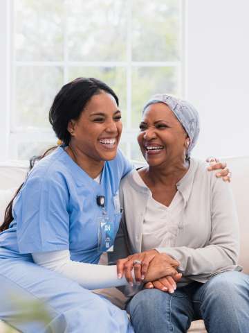 During her visit, the mid adult home health nurse and the senior adult woman with cancer embrace and laugh together.
