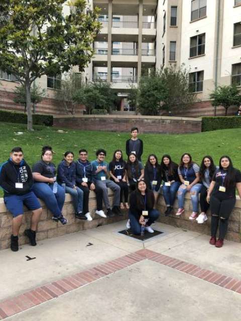 Group of people sitting and standing outdoors in front of a multi-story building, wearing lanyards and casual clothing.