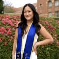 A woman with wavy black hair, wearing a white dress and graduation sash, posing on UCLA campus