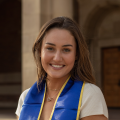 A woman with blonde hair, wearing her UCLA sash, smiling on UCLA campus
