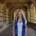 A woman with long blonde hair, wearing a white dress and graduation sash, smiling in a brick hallway on UCLA campus