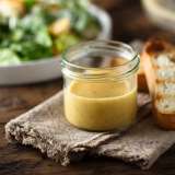 A jar of mustard dressing beside toasted bread on a wooden surface, with a salad in the background.