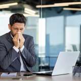 A man with allergies sneezes while at his work desk, covering his nose with a tissue.