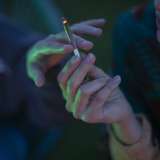 Person holding a cannabis cigarette, with another hand reaching towards it in dim lighting.