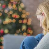 A woman, sitting alone, looks said, with a Christmas tree in the background.