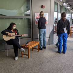 A person wearing a mask sits on a stool and plays a guitar. Two other people stand nearby, looking at a poster on the wall.