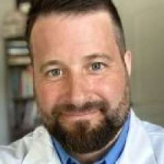 Person wearing a white lab coat over a blue collared shirt, with shelves of books visible in the background.