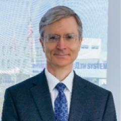 A man with short grey hair, wearing glasses with a suit and tie, posing in front of a building window