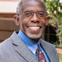 Headshot of Doctor Dale Abel in a grey suit, wearing glasses and standing in front of a brick building with trees to the right.