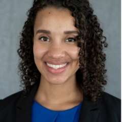 Professional headshot of a person with curly hair, wearing a blue top and black blazer.