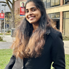 A person with long hair wearing a black coat stands outdoors near a building.
