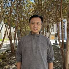 Ke Wang wearing a grey long sleeve shirt with the sleeves rolled up standing in front of some trees